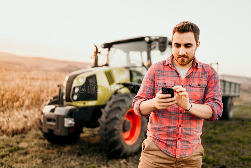 Moderne en landbouwer die gebruikend smartphone in moderne landbouw met tractorachtergrond werken oogsten