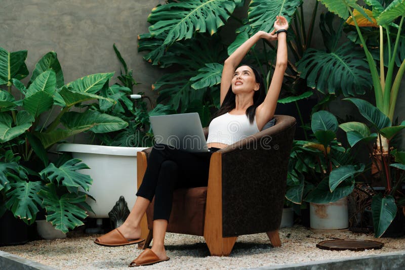 Modern young woman working remotely or relaxing in the solitude of a minimalist architectural concrete style summer exotic plant foliage garden, surrounded by tropical plants. Blithe. Modern young woman working remotely or relaxing in the solitude of a minimalist architectural concrete style summer exotic plant foliage garden, surrounded by tropical plants. Blithe