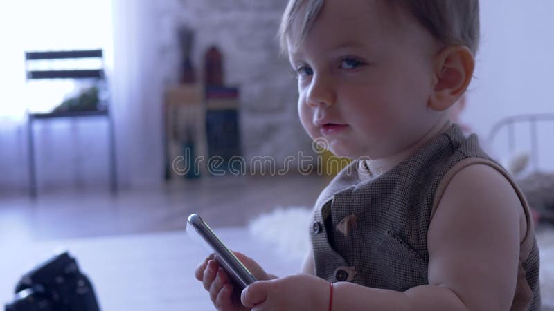 Modernes Baby, das zuhause mit Smartphone auf unfocused Hintergrund spielt