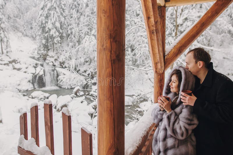 Stylish couple holding hot tea in cups and looking at winter snowy mountains from wooden porch. Happy romantic family with drinks hugging. Space for text. Holiday getaway together. Stylish couple holding hot tea in cups and looking at winter snowy mountains from wooden porch. Happy romantic family with drinks hugging. Space for text. Holiday getaway together.