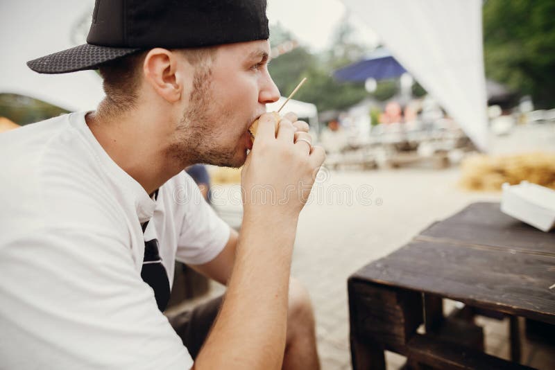 Modieuze hipstermens die heerlijke veganisthamburger eten bij het festival van het straatvoedsel Hongerige mens het bijten hambur stock foto Modieuze hipstermens die heerlijke veganisthamburger eten bij het festival van het straatvoedsel Hongerige mens het bijten hambur stock foto