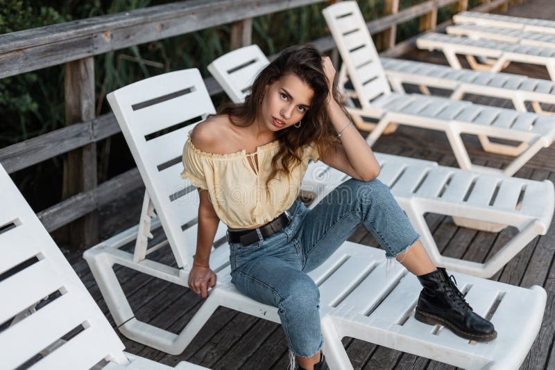 Modna, śliczna, kręcona kobieta hipster w żółtym letnim bluzce modne niebieskie dżinsy z skórzanymi butami spoczywającymi na plaży fotografia royalty free