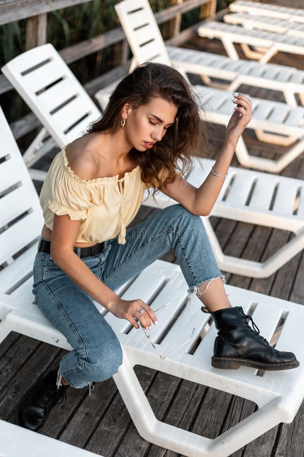 Modny hipster piękna kobieta w letnim dżinsowym ubraniu z butami i spoczywać na lodzie na plaży na drewnianym molo zdjęcia stock