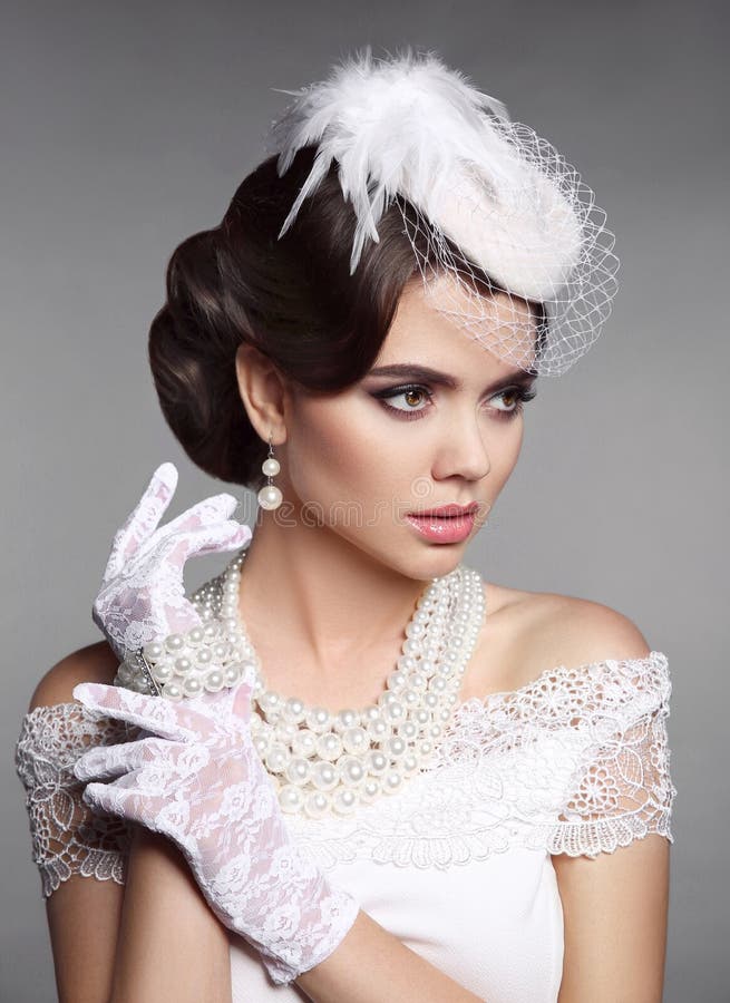 Fashion Retro elegant woman portrait. Wedding hairstyle. Brunette bride model present white hat, gloves and pearls jewelry accessories over gray studio background. Fashion Retro elegant woman portrait. Wedding hairstyle. Brunette bride model present white hat, gloves and pearls jewelry accessories over gray studio background.