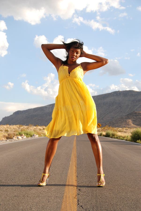 Full length shot of a beautiful African American woman wearing a yellow summer dress standing in the middle of the road in the desert. Full length shot of a beautiful African American woman wearing a yellow summer dress standing in the middle of the road in the desert.