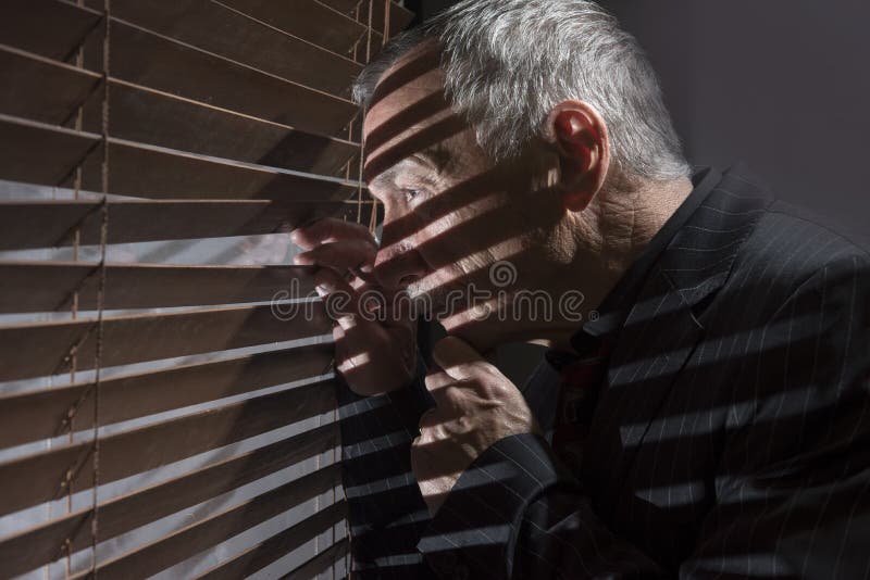 Mogen man som ser ut ur ett fönster med rullgardiner som gjuter skuggor fotografering för bildbyråer