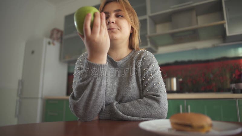 Molliges Mädchen denkt, dass sie einen geschmackvollen Hamburger oder einen saftigen grünen Apfel essen sollte Schwierige Wahl zw