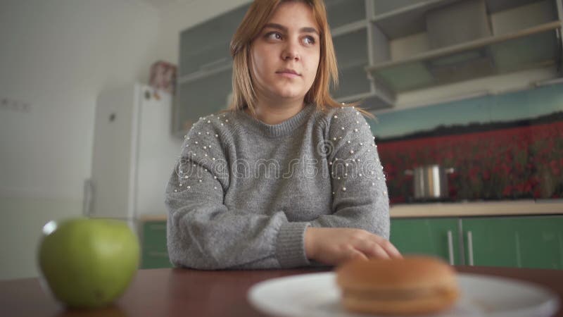 Molliges Mädchen des Porträts denkt, dass sie einen geschmackvollen Hamburger oder einen saftigen grünen Apfel essen sollte Schwi