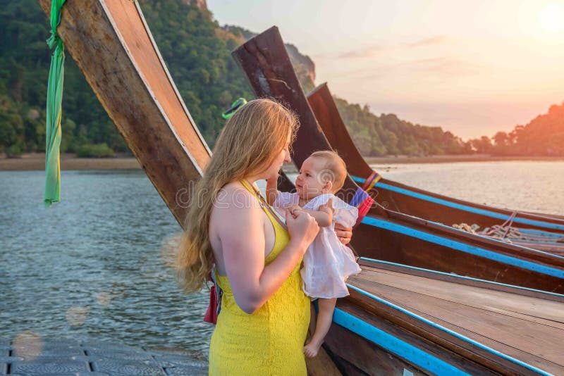 Mom and baby are standing on a pontoon pier next to wooden boats. The kid is studying mom with interest. Early morning, sunrise on stock photography Mom and baby are standing on a pontoon pier next to wooden boats. The kid is studying mom with interest. Early morning, sunrise on stock photography