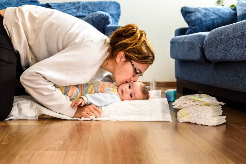 Mom changing the diaper to her daughter, on the floor of her living room and giving her kisses.