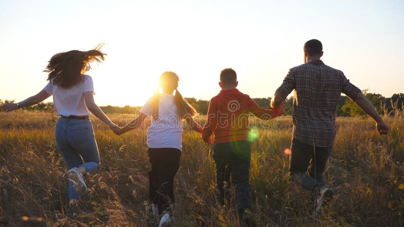Mom and dad with two kids holding hands of each other and running through grass field at sunset. Happy parents with