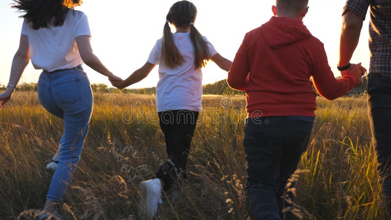 Mom and dad with two kids holding hands of each other and running through grass field at sunset. Happy parents with