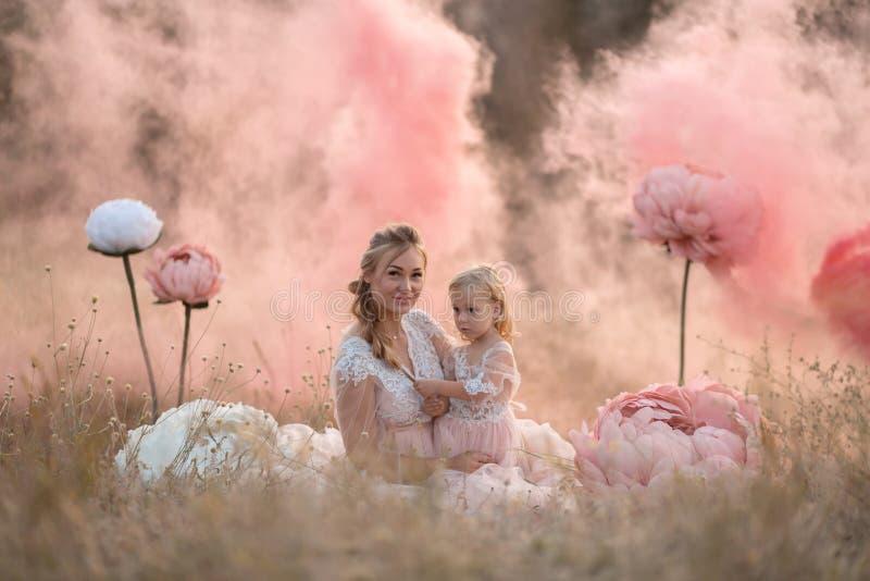 Mom and daughter in pink fairy-tale dresses are sitting in a field surrounded by Big pink decorative flowers stock photo Mom and daughter in pink fairy-tale dresses are sitting in a field surrounded by Big pink decorative flowers stock photo