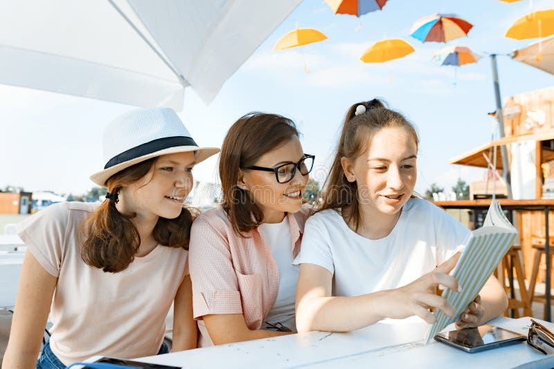 Mom and daughters teenagers have fun, talking, look and read funny book. Communication of the parent and children of adolescents. Sit in a street cafe, sunny summer day.