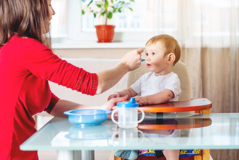 Mom feeding the baby holding out her hand with a spoon of food in the kitchen. Emotions of a child while eating stock photo Mom feeding the baby holding out her hand with a spoon of food in the kitchen. Emotions of a child while eating stock photo