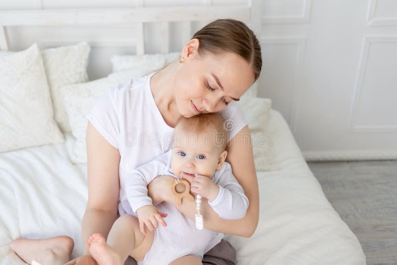 Mom holds baby in her arms with a rodent on a white bed with cotton bedding at home, teether