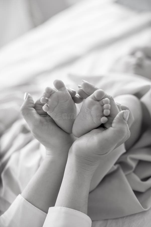 Mom`s hands are holding little cute legs of a newborn baby at home on a white bed