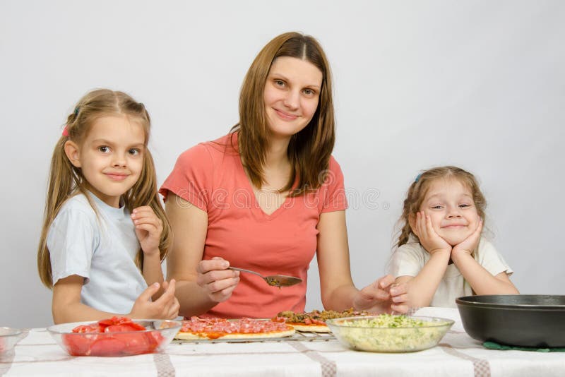 Momia con dos niñas que se sientan en la tabla de cocina que prepara una pizza imagen de archivo libre de regalías Momia con dos niñas que se sientan en la tabla de cocina que prepara una pizza imagen de archivo libre de regalías