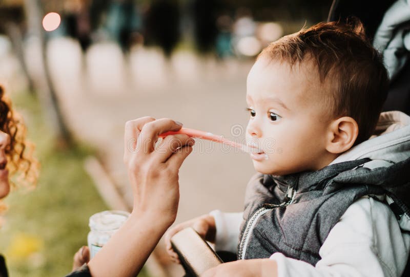 Mommy and child, eating and parenthood - mother with puree and spoon feeding little baby in park in stroller