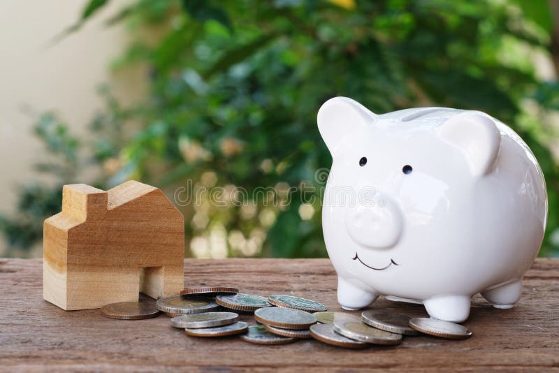 Money for housing. Wooden house model, piggy bank and pile of coins with greenery background