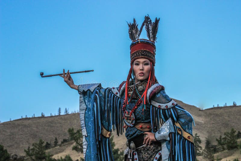 Mongoloid женщина в костюме шамана и ведьмы танцует и курит труба на фоне гор стоковая фотография Mongoloid женщина в костюме шамана и ведьмы танцует и курит труба на фоне гор стоковая фотография