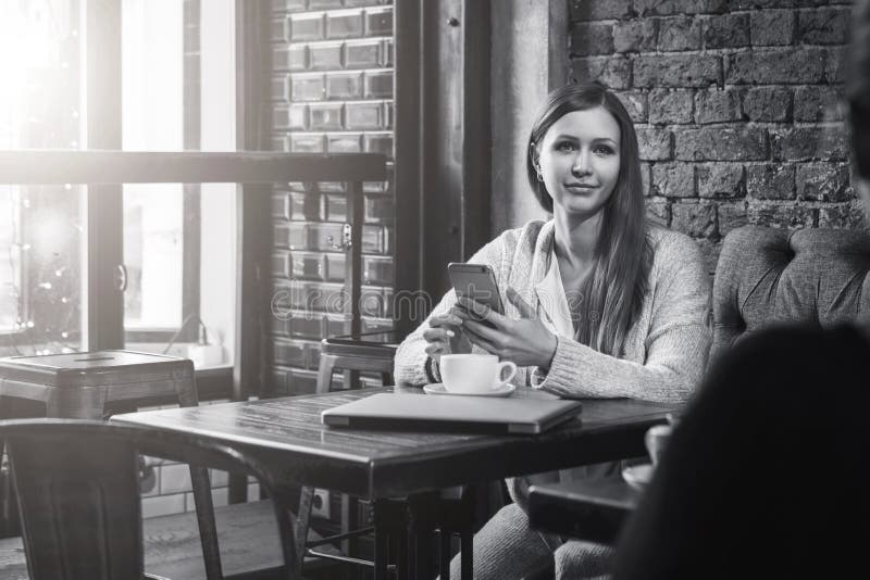 Monokromen avbildar Den unga kvinnan sitter på tabellen i kafé och brukssmartphone På tabellen är koppen kaffe och den stängda an arkivfoton
