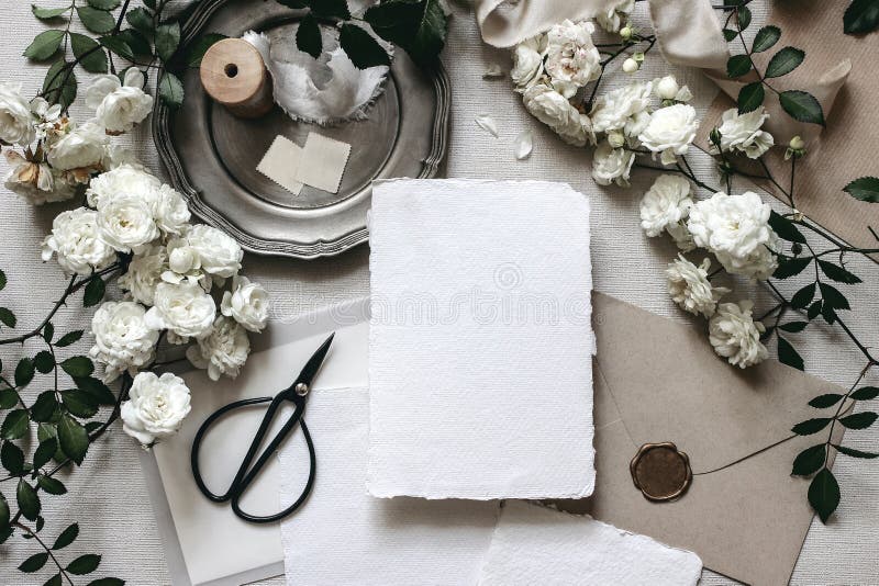 Moody wedding table mockup scene. Stationery composition with fading white rose flowers, silver plate, black scissors