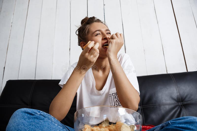 Mooi binnenlands meisje die spaanders eten, lettend op TV, die bij bank zitten royalty-vrije stock foto Mooi binnenlands meisje die spaanders eten, lettend op TV, die bij bank zitten royalty-vrije stock foto