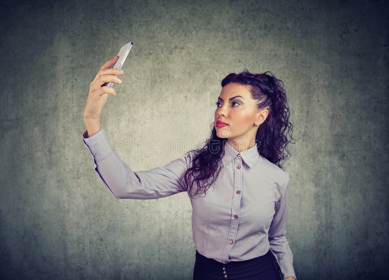 Mooi brunette in formele uitrusting gebruikend smartphone en nemend selfie op grijze achtergrond