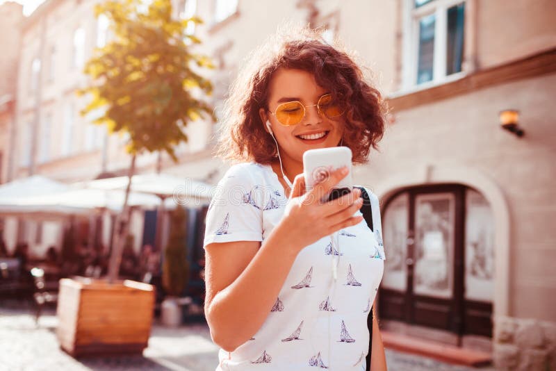 Mooi jong meisje gebruikend slimme telefoon en luisterend aan de muziek die op straat lopen royalty-vrije stock foto's Mooi jong meisje gebruikend slimme telefoon en luisterend aan de muziek die op straat lopen royalty-vrije stock foto's