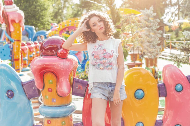 Mooi jong vrolijk meisje met krullend haar in denimborrels en witte T-shirt bij een pretpark bij zonsondergang heldere zon royalty-vrije stock afbeeldingen Mooi jong vrolijk meisje met krullend haar in denimborrels en witte T-shirt bij een pretpark bij zonsondergang heldere zon royalty-vrije stock afbeeldingen