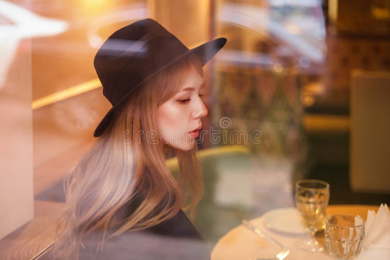 Mooi jong vrouwenblonde in hoed bij lijst in een restaurant door glas Bezinningen stock afbeelding Mooi jong vrouwenblonde in hoed bij lijst in een restaurant door glas Bezinningen stock afbeelding