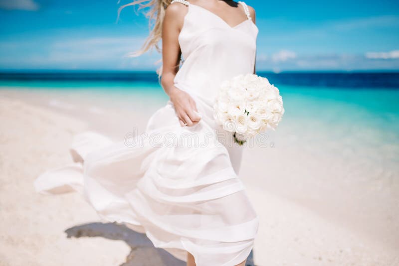 Beautiful blond bride in white wedding dress with white wedding bouquet stand on sea shore. Tropical turquois sea on the background. Summer vacation concept. Beautiful blond bride in white wedding dress with white wedding bouquet stand on sea shore. Tropical turquois sea on the background. Summer vacation concept.