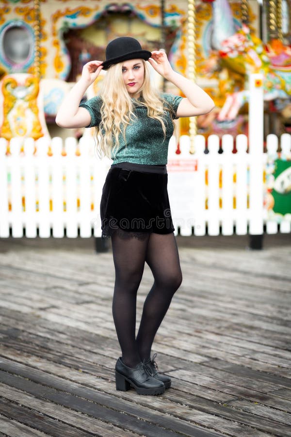 Portrait of a beautiful confident blond blue eyed teenage girl with long hair wearing a bowler hat, green glittery t-shirt dark red lipstick standing alone in front of a fairground ride with a white fence. Portrait of a beautiful confident blond blue eyed teenage girl with long hair wearing a bowler hat, green glittery t-shirt dark red lipstick standing alone in front of a fairground ride with a white fence.