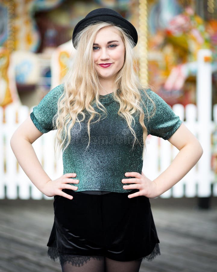 Portrait of a beautiful confident blond blue eyed teenage girl with long hair wearing a bowler hat, green glittery t-shirt dark red lipstick standing alone in front of a fairground ride with a white fence. Portrait of a beautiful confident blond blue eyed teenage girl with long hair wearing a bowler hat, green glittery t-shirt dark red lipstick standing alone in front of a fairground ride with a white fence.