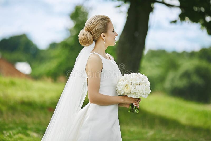Mooie en elegante jonge blondevrouw in modieuze huwelijkskleding met boeket van witte bloemen in haar handen die in openlucht ste royalty-vrije stock afbeelding Mooie en elegante jonge blondevrouw in modieuze huwelijkskleding met boeket van witte bloemen in haar handen die in openlucht ste royalty-vrije stock afbeelding