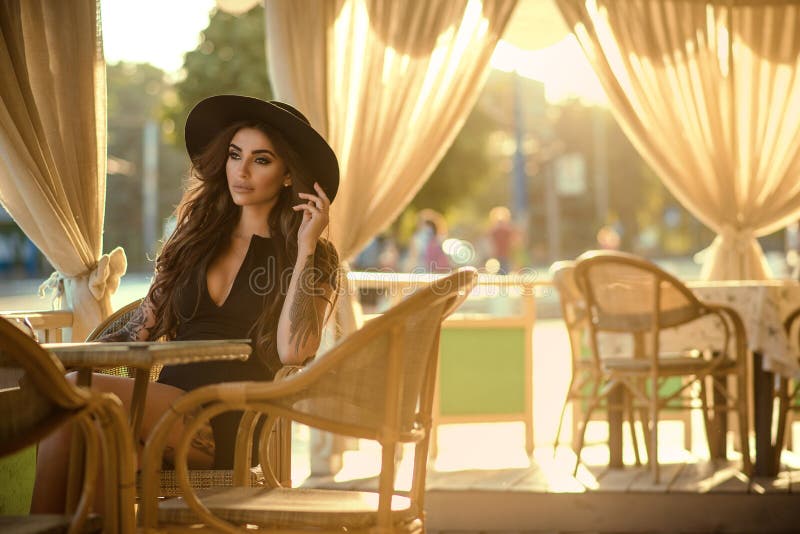 Beautiful glam tattooed brunette in little black dress and trendy fedora hat sitting in the nice open air summer restaurant in the city at sunset and looking aside with sadness in her eyes. Copy-space. Beautiful glam tattooed brunette in little black dress and trendy fedora hat sitting in the nice open air summer restaurant in the city at sunset and looking aside with sadness in her eyes. Copy-space