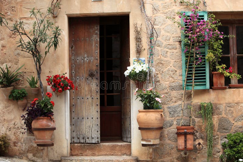 Pretty entrance doorway to a rural house surrounded by colourful flowering plants in terracotta urns and pots and a window with a blue painted shutter. Pretty entrance doorway to a rural house surrounded by colourful flowering plants in terracotta urns and pots and a window with a blue painted shutter