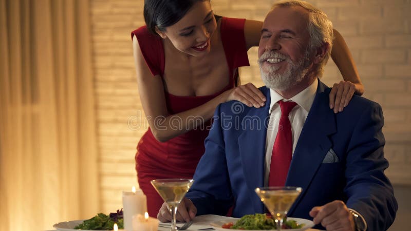 Mooie jonge dame die schouders die van oude rijke man masseren, op datum flirten royalty-vrije stock afbeelding Mooie jonge dame die schouders die van oude rijke man masseren, op datum flirten royalty-vrije stock afbeelding