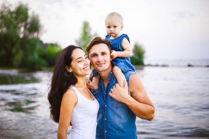 Mooie jonge familie op vakantie met baby De vader houdt het blondemeisje in haar wapens, en de donkerbruine moeder van ` s koeste stock foto Mooie jonge familie op vakantie met baby De vader houdt het blondemeisje in haar wapens, en de donkerbruine moeder van ` s koeste stock foto