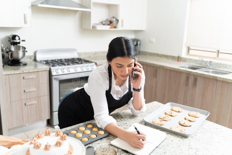 Gorgeous female baker writing order in diary while talking to customer through mobile phone at kitchen counter. Gorgeous female baker writing order in diary while talking to customer through mobile phone at kitchen counter