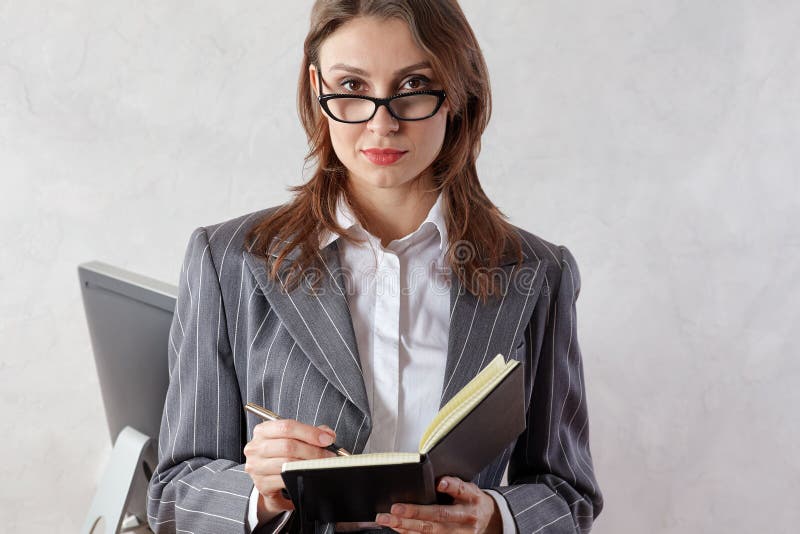 Mooie jonge professionele donkerbruine vrouw in bureau met oogglazen, die in een stootkussen, met zekere uitdrukking schrijven royalty-vrije stock afbeeldingen Mooie jonge professionele donkerbruine vrouw in bureau met oogglazen, die in een stootkussen, met zekere uitdrukking schrijven royalty-vrije stock afbeeldingen