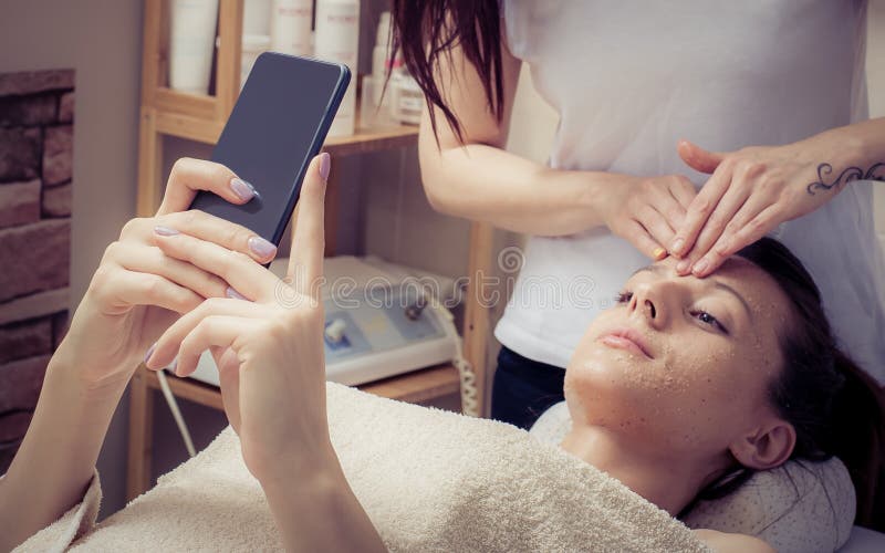 Mooie jonge vrouw met gezichtsmasker die smartphone in schoonheidssalon gebruiken stock foto Mooie jonge vrouw met gezichtsmasker die smartphone in schoonheidssalon gebruiken stock foto