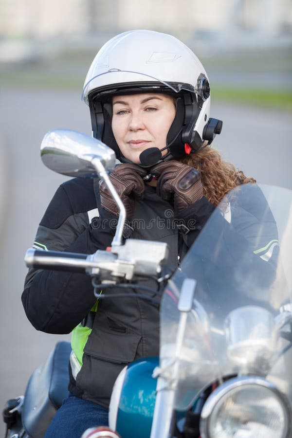 Mooie motorrijder Kaukasische vrouw die op witte helm proberen terwijl het zitten op fiets en het bekijken achterspiegel stock afbeelding Mooie motorrijder Kaukasische vrouw die op witte helm proberen terwijl het zitten op fiets en het bekijken achterspiegel stock afbeelding