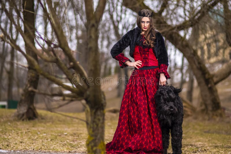 Mooie Prinses in Rode Kleding en het Zwarte Bontjasje Stellen in Kroon samen met Haar Hond in Forest During Early Spring Art