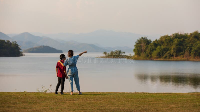 Mor och son som står bredvid stora sjön och ser bergsvyn i bakgrunden när mamma pekar finger åt skogen. familjeidé
