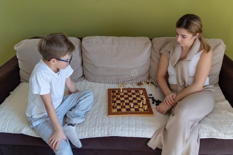 Mor och son spelar schack på en beige soffa. En kvinna och en pojke sitter på en beige soffa framför en grön vägg. En kvinna och en pojke sitter på en beige-soffa framför en grön vägg som koncentrerar sig på ett schackspel som placeras mellan dem med spridda schackbitar runt bordet.