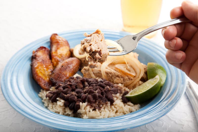 Closeup of a piece of marinated roast pork on a fork, about to be eaten. Plate of traditional Cuban food in the background, including black beans and rice, and fried plantains. Closeup of a piece of marinated roast pork on a fork, about to be eaten. Plate of traditional Cuban food in the background, including black beans and rice, and fried plantains.