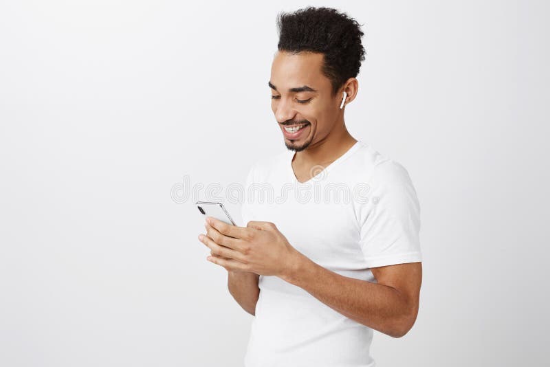 Mordern guy choose new devices. Attractive dark-skinned urban citizen in white t-shirt, listening music in wireless earbuds, holding smartphone, smiling at screen while messaging or watching video. Technology concept