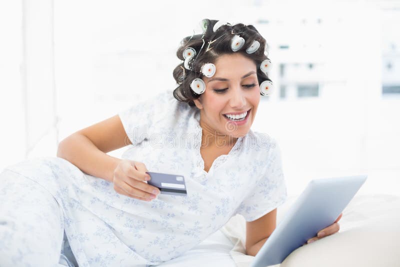 Morena de sorriso nos rolos do cabelo que encontram-se em sua cama usando seu tabl imagem de stock Morena de sorriso nos rolos do cabelo que encontram-se em sua cama usando seu tabl imagem de stock